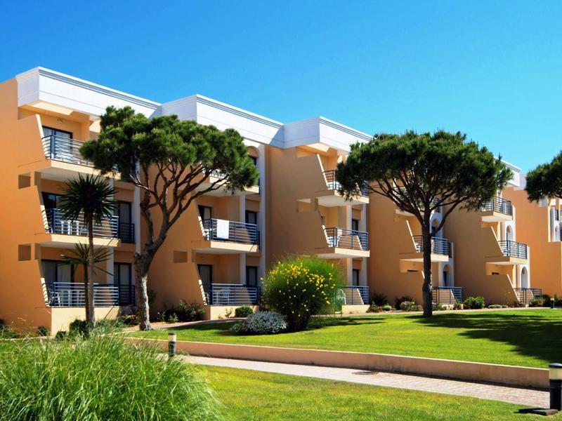 Mehrstöckige, beige Wohnungen mit Balkonen, umgeben von grünen Bäumen und gepflegtem Rasen.