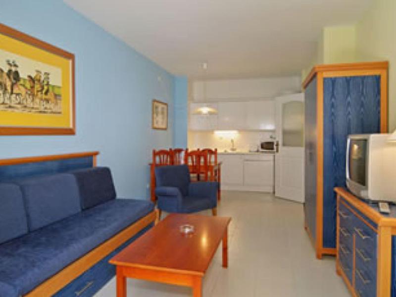 Wohnzimmer mit blauer Couch, Holztisch, Sessel, Schrank und angrenzender Küche in hellen Farben.