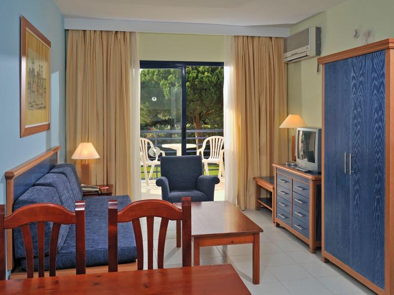 Wohnzimmer mit blauem Sofa, Sessel, Holzmöbeln, Fernseher und Zugang zu Balkon mit grüner Sicht.