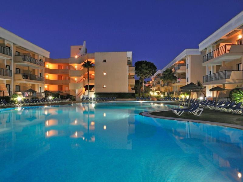 Ein beleuchteter Pool umgeben von mehrstöckigen weißen Wohngebäuden in der Abenddämmerung.