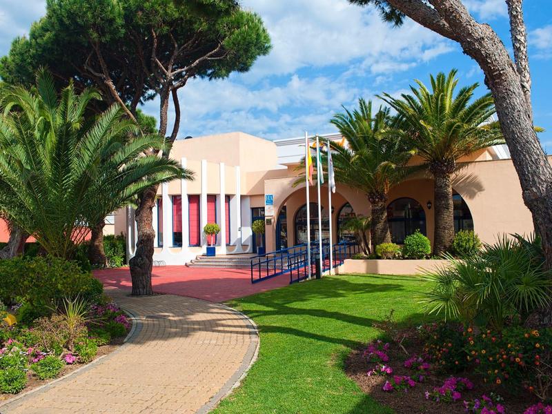 Hellrosa Gebäude mit Säulen, Palmen und bunten Blumen in gepflegtem Garten unter blauem Himmel.