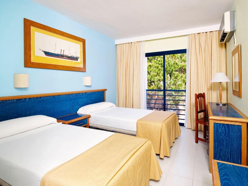 Hellblaues Hotelzimmer mit zwei Einzelbetten, Holzmöbeln und Balkon mit grünem Ausblick.