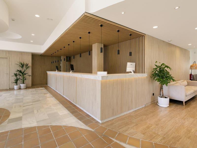 Heller Empfangsbereich mit langem Holztresen, Sessel, Pflanzen und moderner Deckenbeleuchtung.