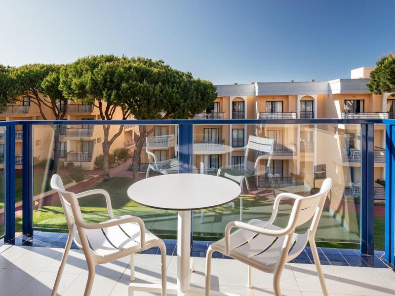 Balkon mit Tisch und Stühlen mit Blick auf einen Hotelgarten und Gebäude bei klarem Himmel.