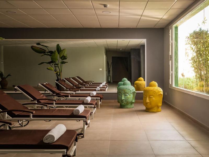 Ruhebereich mit braunen Liegen, großen Pflanzen und bunten Skulpturen vor einem Fenster.