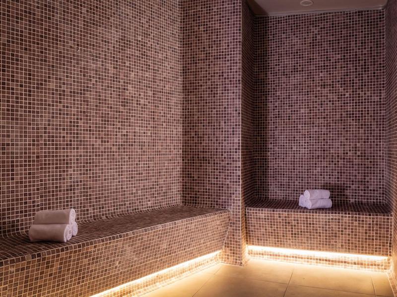 Dampfbad mit braunen Mosaikfliesen, Sitzbänken und zwei aufgerollten weißen Handtüchern.