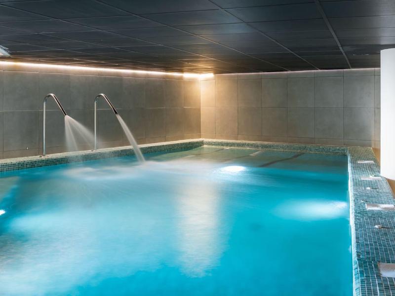 Modernes Hallenbad mit blauem Wasser, Wasserdüsen und grauen Fliesenwänden, diffuse Beleuchtung.