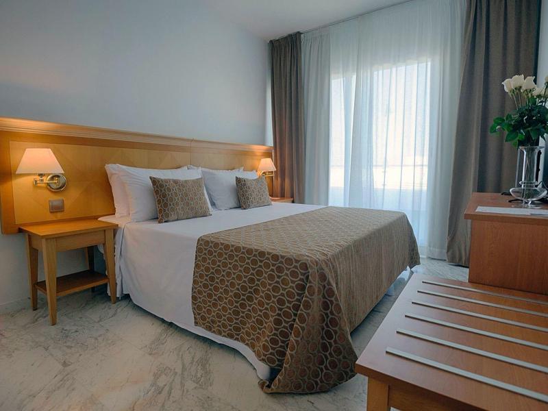 Hell eingerichtetes Schlafzimmer mit großem Bett, Nachttisch, Gardinen und Holzmöbeln.