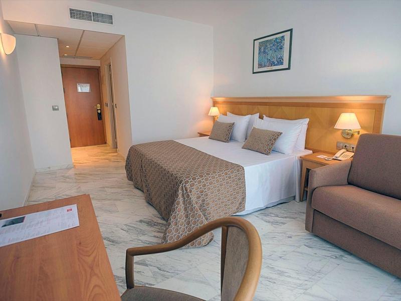 Hell eingerichtetes Hotelzimmer mit Doppelbett, Sofa, Holzschreibtisch und moderner Beleuchtung.