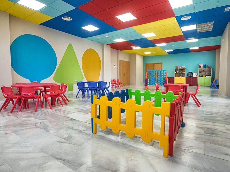 Bunter Spielraum mit kleinen Tischen, Stühlen und farbigem Zaun auf grauem Boden.