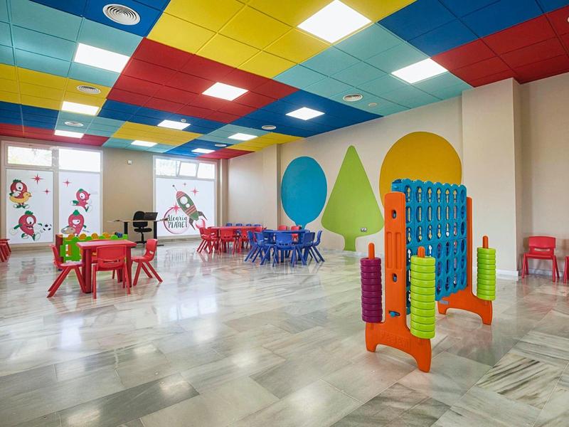 Bunter, großzügiger Kindergartenraum mit Tischen, Stühlen und Spielgeräten auf Marmorfußboden.