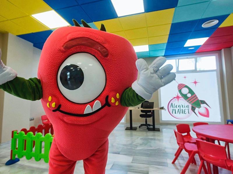 Roter Maskottchen-Charakter mit einem großen Auge in einem bunten Raum mit Tischen und Stühlen.