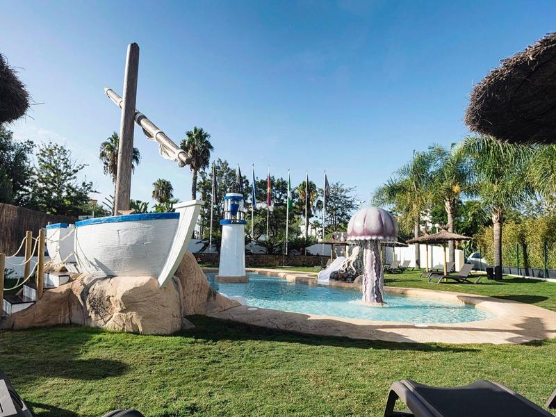 Pool mit Wasserpilz und Boot-Deko, umgeben von Palmen und Sonnenschirmen unter klarem Himmel.