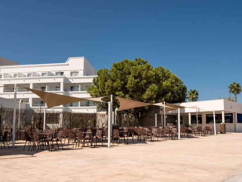 Modernes Gebäude mit Terrassenmöbeln, Sonnenschirmen und Bäumen unter klarem blauem Himmel.