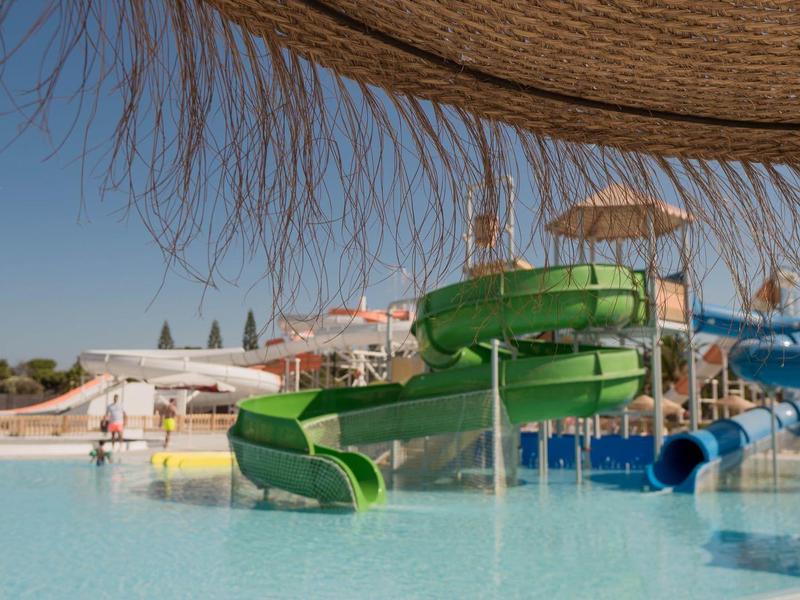 Wasserpark mit grüner und blauer Rutsche, großem Pool und Sonnenschirm im Vordergrund.