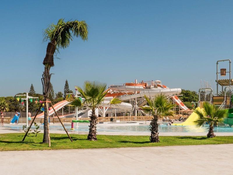 Freizeitpark mit Rutschen, Pool, Palmen und Personen bei sonnigem Wetter unter blauem Himmel.