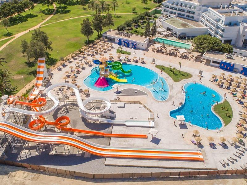 Buntes Schwimmbad mit mehreren Wasserrutschen, Liegestühlen und angrenzendem Hotel in grüner Umgebung.