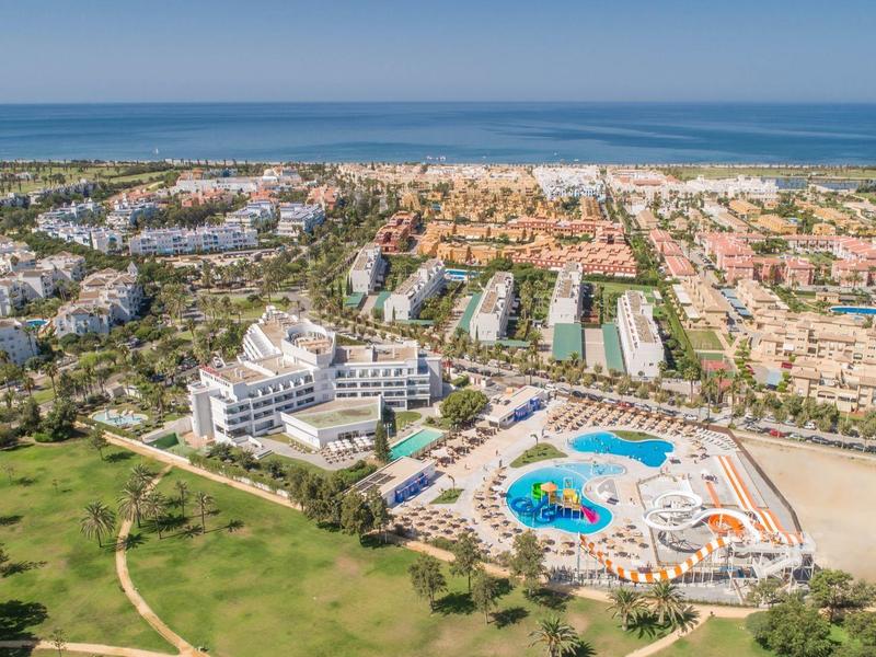 Luftaufnahme eines großen Resorts mit Pool, weißen Gebäuden und Strand am Horizont.