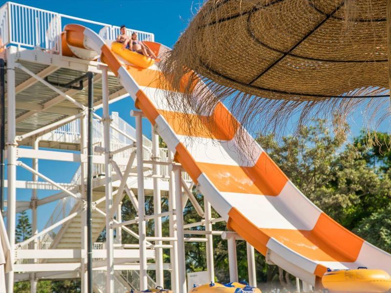 Wasserrutsche in Orange-Weiß mit Metallgestell und Strohhut-Schirm im Vordergrund an sonnigem Tag.