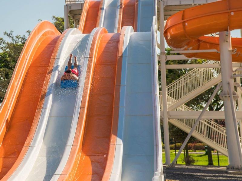 Drei bunte Wasserrutschen mit einem Fahrer auf der blauen, umgeben von Bäumen und Baugerüst.