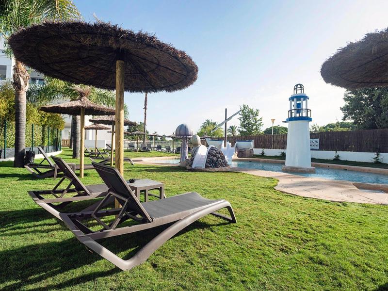 Liegestühle mit Strohdächern stehen auf grünem Rasen neben einem Pool mit kleinem Leuchtturm.