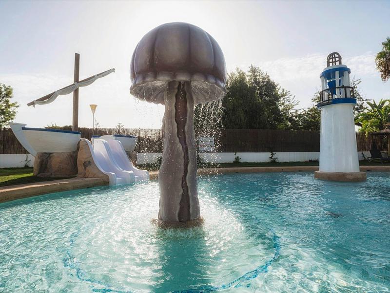 Schwimmbecken mit Wasserrutsche, Pilz-Wasserspiel und Leuchtturm-Deko bei Sonnenschein.