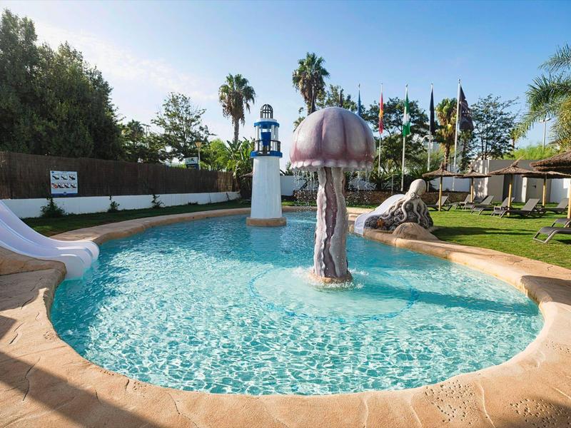 Runder Kinderpool mit Wasserspiel in Pilzform, Wasserrutsche und Palmen im Hintergrund.