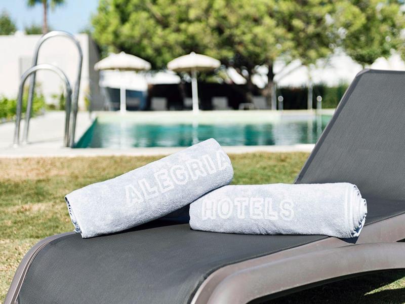 Zwei weiße Handtücher liegen auf einer grauen Liege neben einem Pool mit grüner Wiese und Bäumen.