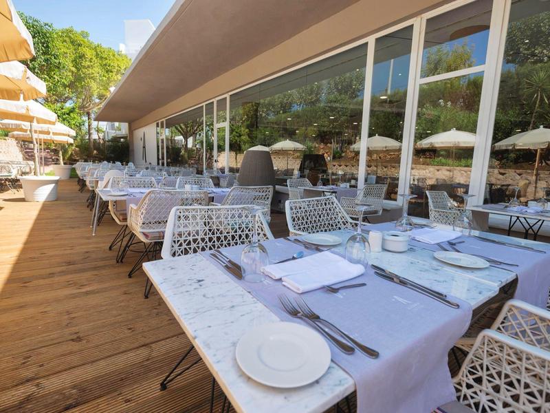 Terrassenrestaurant mit weißen Tischen, Stühlen und Sonnenschirmen bei klarem Himmel.