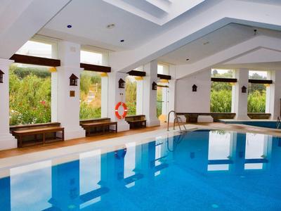 Hallenbad mit klarem blauem Wasser, weißen Wänden, Holzsitzen und großen Fensterfronten.