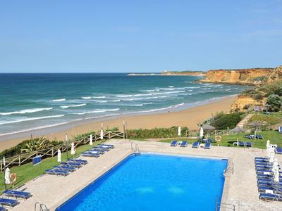 Ein Hotelpool mit blauen Liegestühlen neben einem Sandstrand und ruhigem Meer unter klarem Himmel.