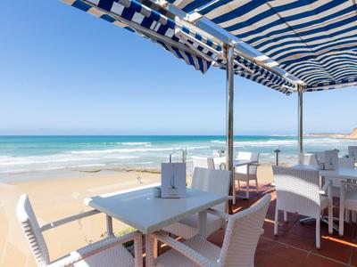 Strandterrasse mit weißen Tischen und Stühlen unter blau-weiß gestreifter Markise am Meer