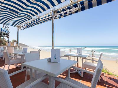 Terrassenbereich mit weißen Stühlen und Tischen unter blau-weiß gestreiftem Sonnenschutz am Meer.