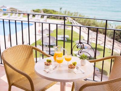 Balkontisch mit zwei Stühlen, Saftgläsern und kleinen Speisen, Meer und Pool im Hintergrund.