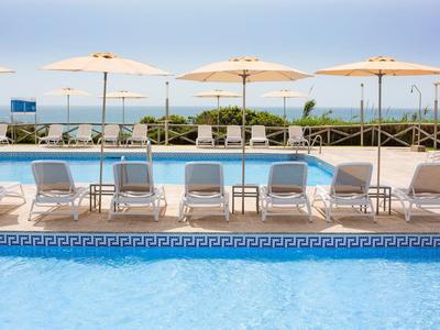 Blauer Pool mit weißen Liegestühlen und Sonnenschirmen, dahinter Meer und blauer Himmel.