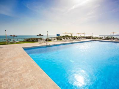 Großer, klarer Pool mit blauem Wasser, umgeben von hellem Steinboden und Liegestühlen am Meer.