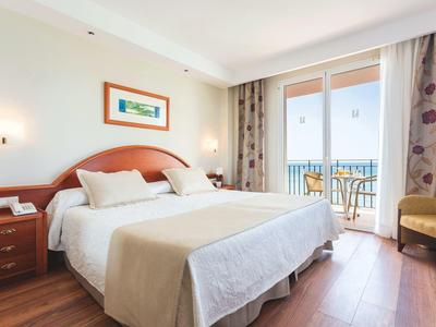Helles Hotelzimmer mit Doppelbett, Holzboden, Nachttisch, Balkon und Blick aufs Meer.