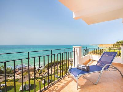 Meerblick von Balkon mit blauem Liegestuhl, hellbraunem Boden und schwarzem Geländer bei Sonnenschein.