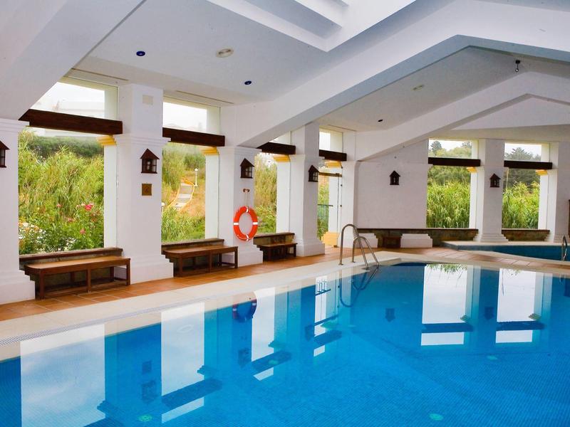 Hallenbad mit klarem blauem Wasser, weißen Wänden, Holzsitzen und großen Fensterfronten.
