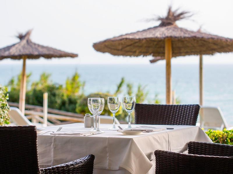 Terrassentisch mit weißer Decke, Weingläsern, braunen Stühlen und Palmen-Sonnenschirmen am Meer.
