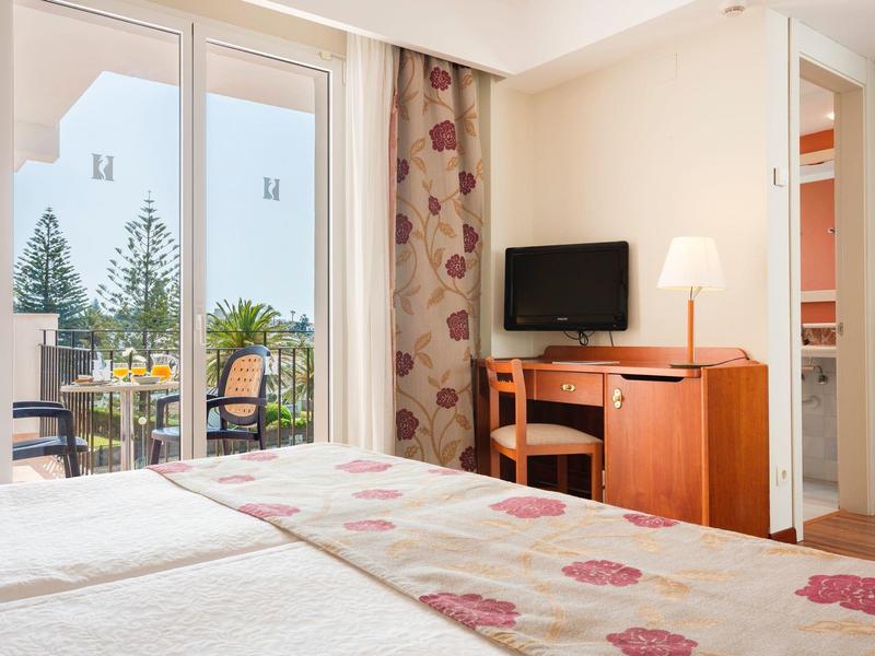 Helles Hotelzimmer mit Doppelbett, Fernseher, Schreibtisch, Lampe und Blick auf Balkon mit Bäumen.