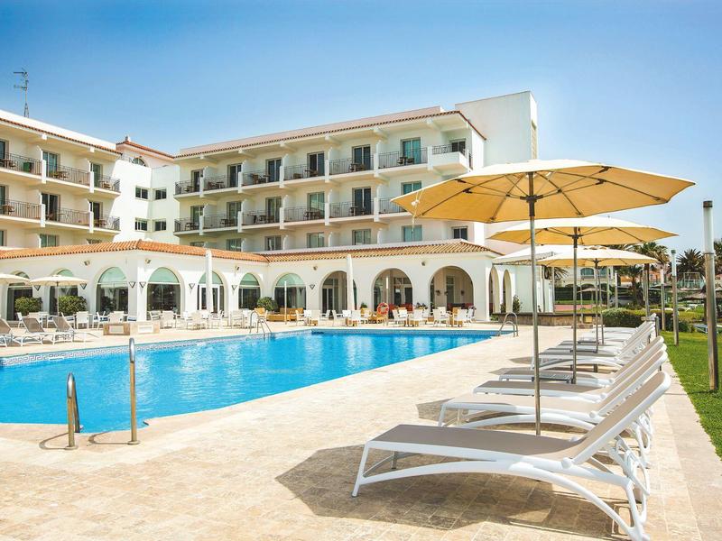 Moderner Hotelpool mit Liegestühlen und gelben Sonnenschirmen neben einem dreistöckigen Gebäude.