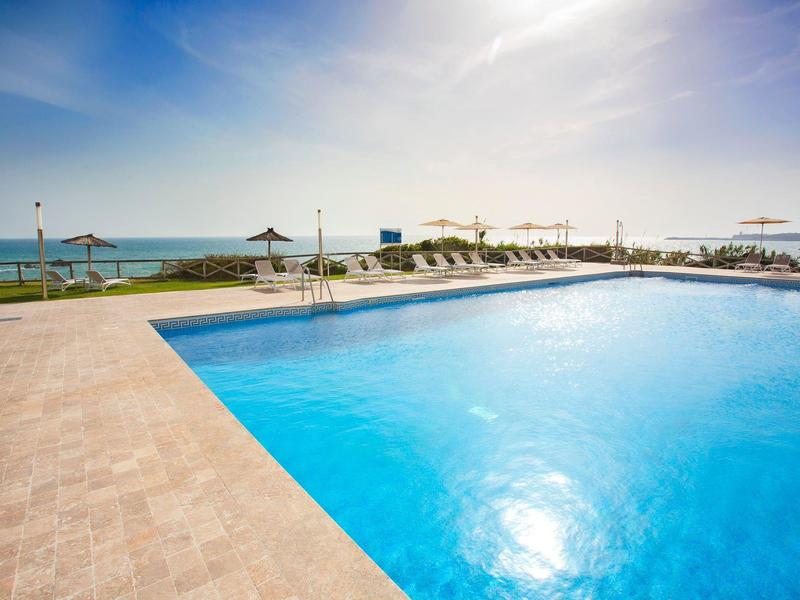 Großer, klarer Pool mit blauem Wasser, umgeben von hellem Steinboden und Liegestühlen am Meer.