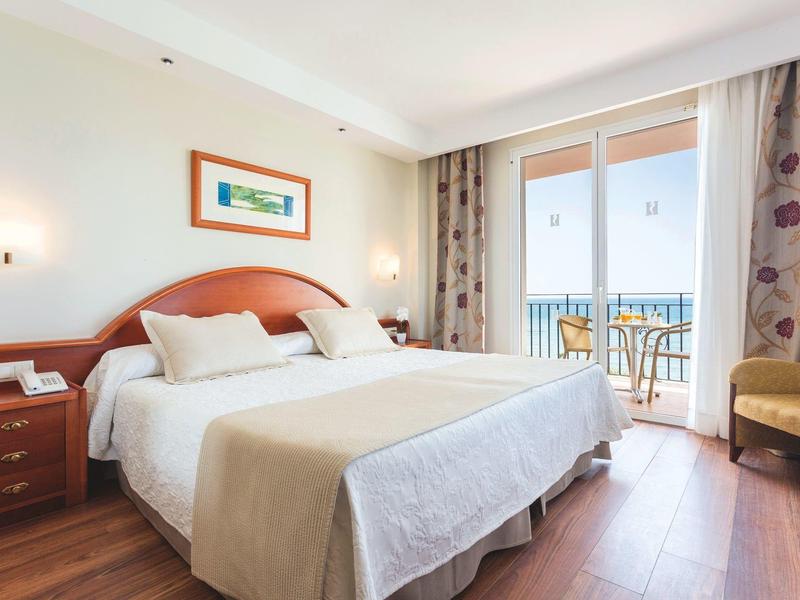 Helles Hotelzimmer mit Doppelbett, Holzboden, Nachttisch, Balkon und Blick aufs Meer.