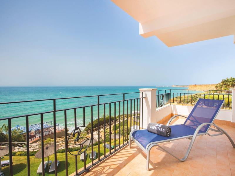 Meerblick von Balkon mit blauem Liegestuhl, hellbraunem Boden und schwarzem Geländer bei Sonnenschein.