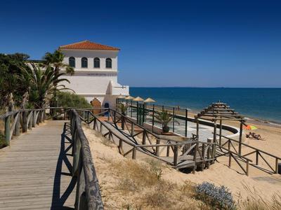 Holzsteg führt zu weißem Haus am Sandstrand mit Palmen und blauem Himmel am Meer.