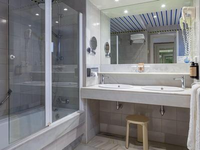 Jasna łazienka hotelowa z dużym szklanym prysznicem, długą umywalką i stołkiem.