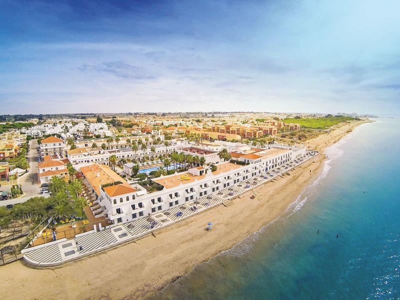 Vista aerea di una spiaggia con un complesso alberghiero lungo la costa e mare calmo sotto un cielo nuvoloso.