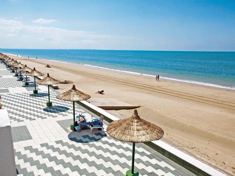 Spiaggia ampia con ombrelloni di paglia e passerella a zigzag vicino al mare.