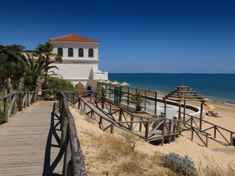 Sentiero in legno verso la spiaggia con edificio bianco dal tetto rosso e mare calmo.